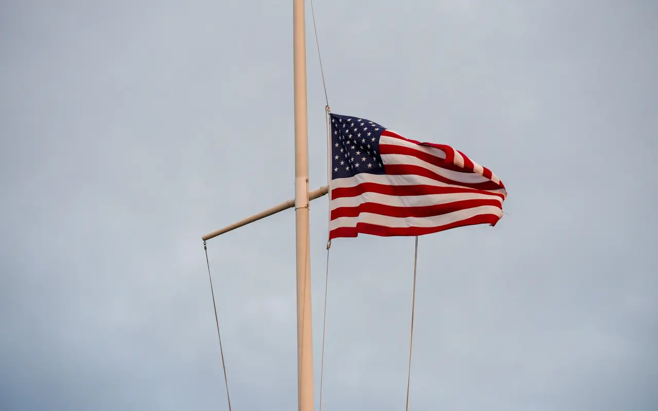 An American flag flying at half-staff on a white yardarm mast against a pale grey sky