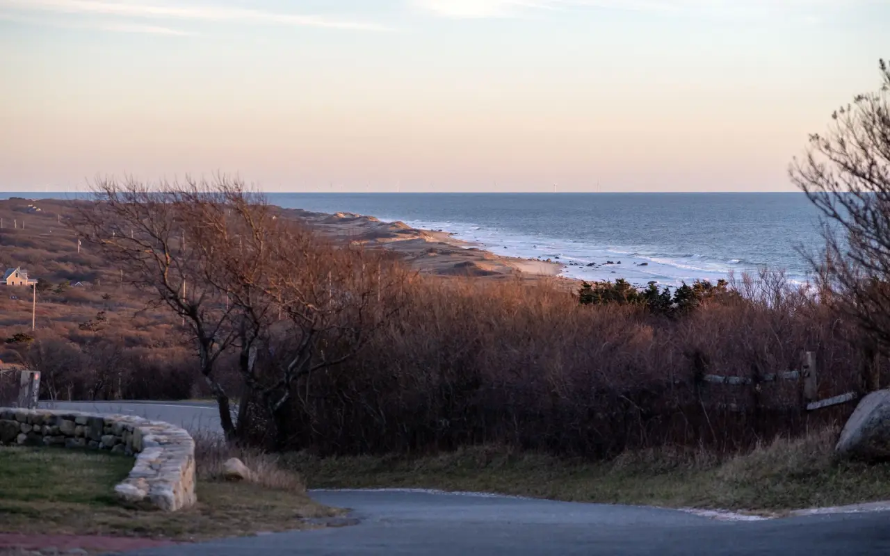 Dusk over the Aquinnah shore: bare scrub on the slope, a stone wall and curving road in front, the ocean and a long beach beyond