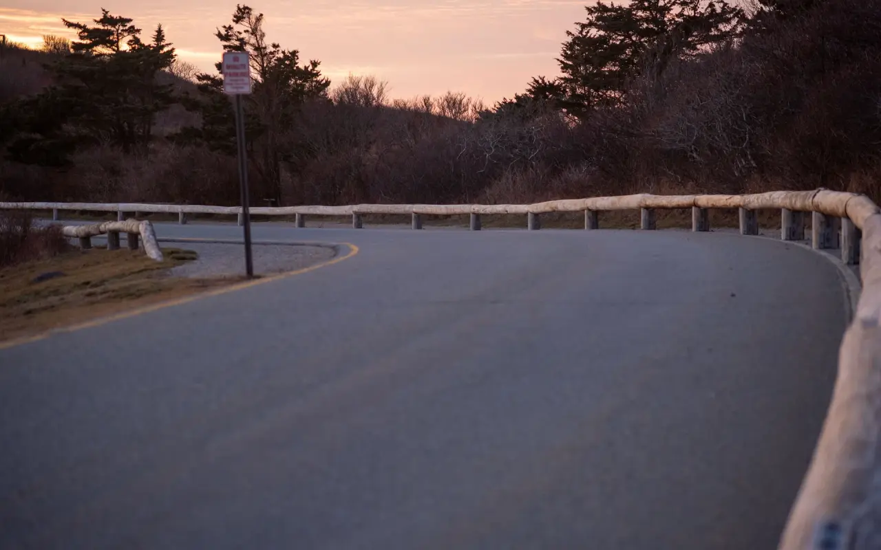 An empty curving road with a metal guard rail and bare trees at dusk in Aquinnah