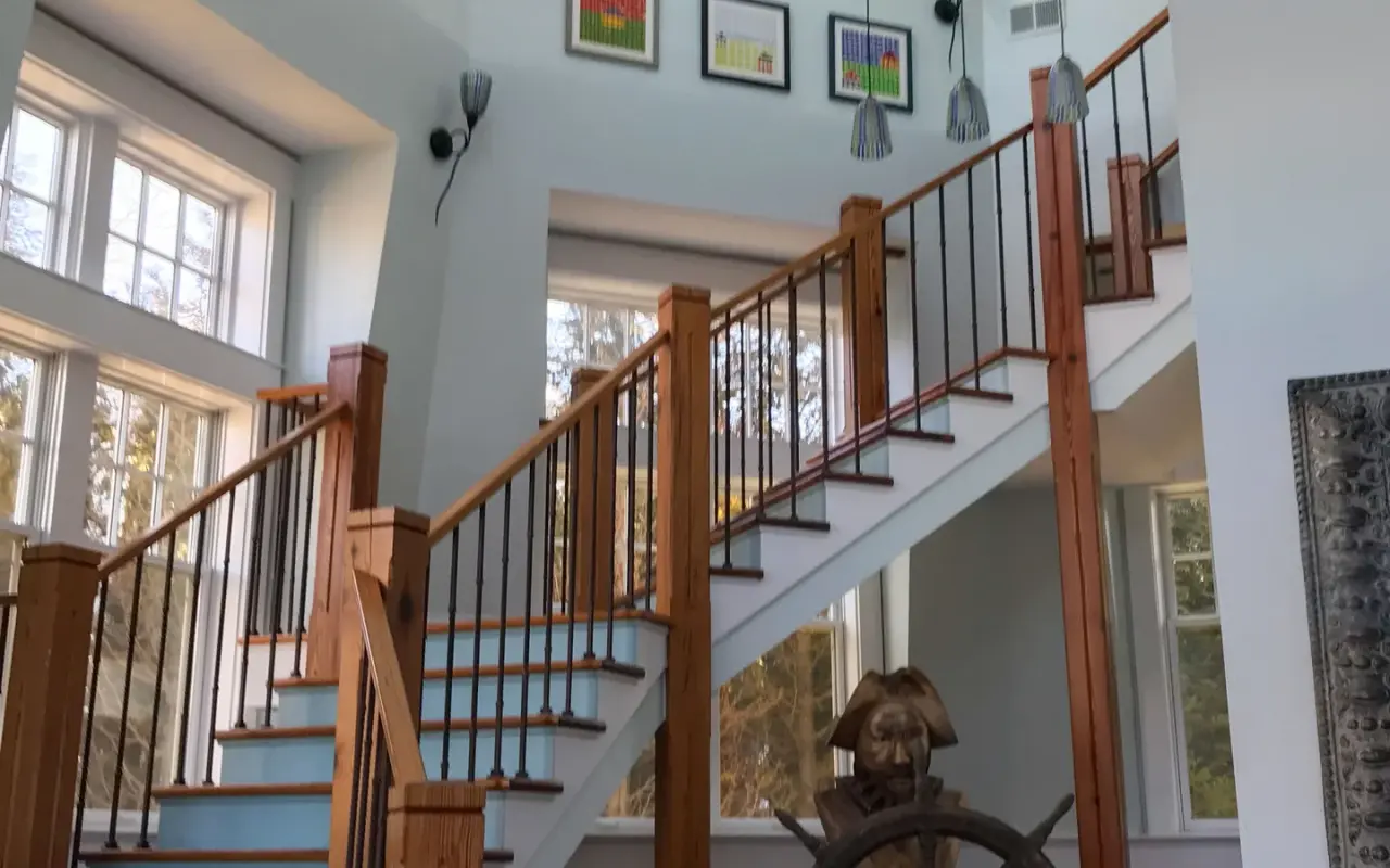 A bright stair hall inside an Edgartown captain's house with white walls, a wooden banister and a ship's wheel