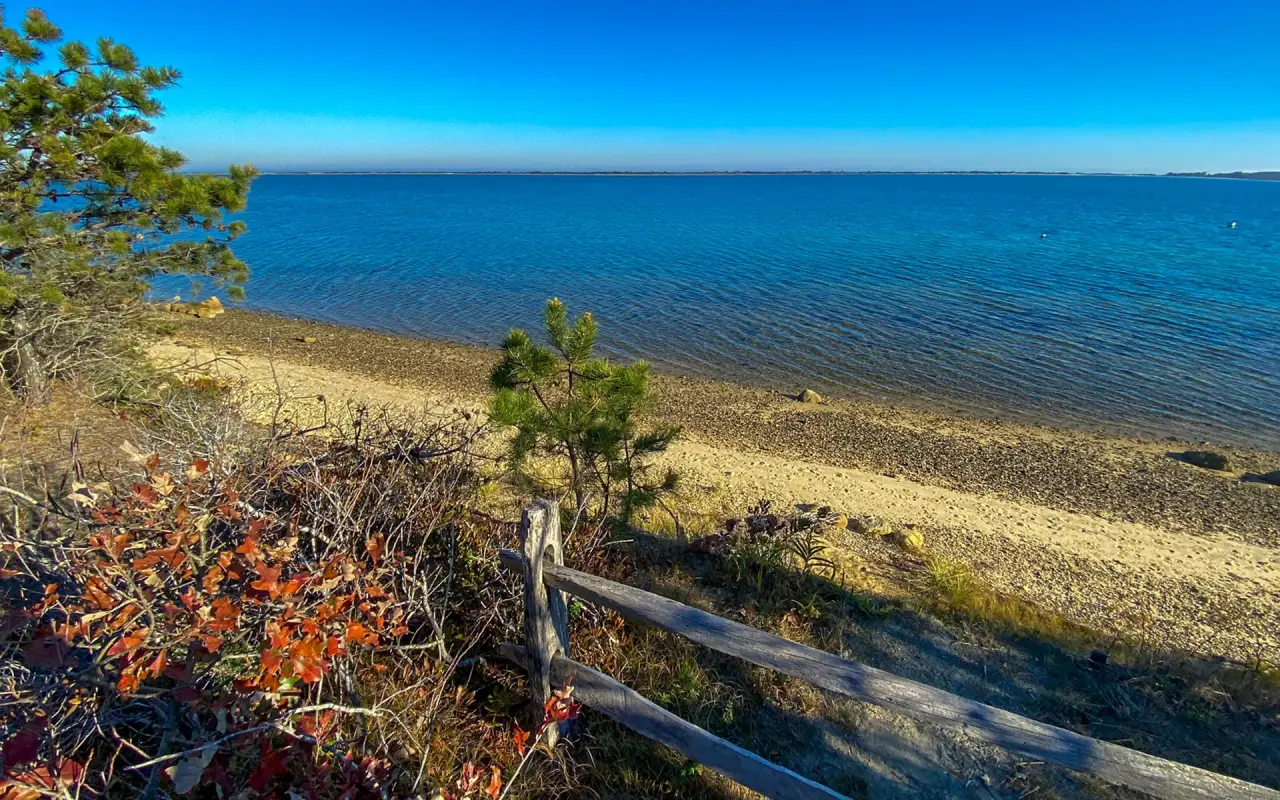 A split-rail fence above a pebbly Chappaquiddick shore with blue water beyond