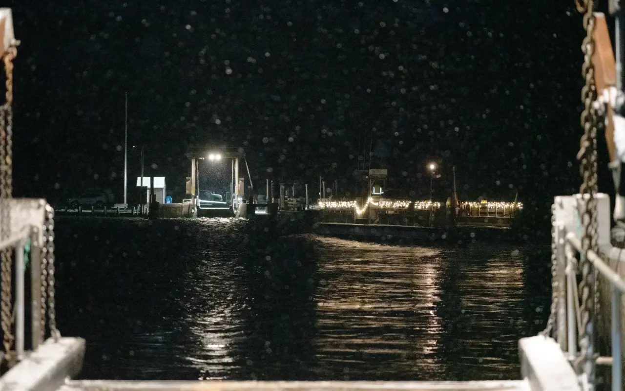 Snow falling at night over Edgartown Harbor seen from the Chappaquiddick ferry ramp with holiday lights on the wharf