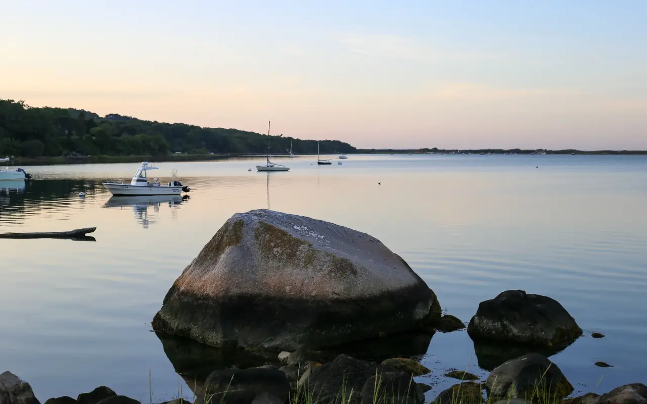A large boulder at the edge of a calm pond at dusk with small boats moored beyond