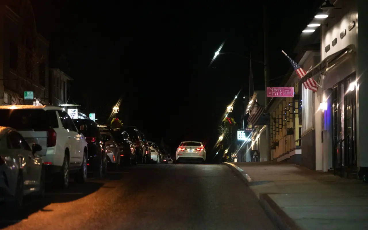 Cars parked along the curb on Circuit Avenue in Oak Bluffs at night, lit shop fronts and an American flag on the right