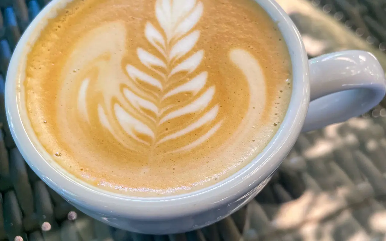 A cup of latte with leaf-pattern foam art on a wicker table in sunlight