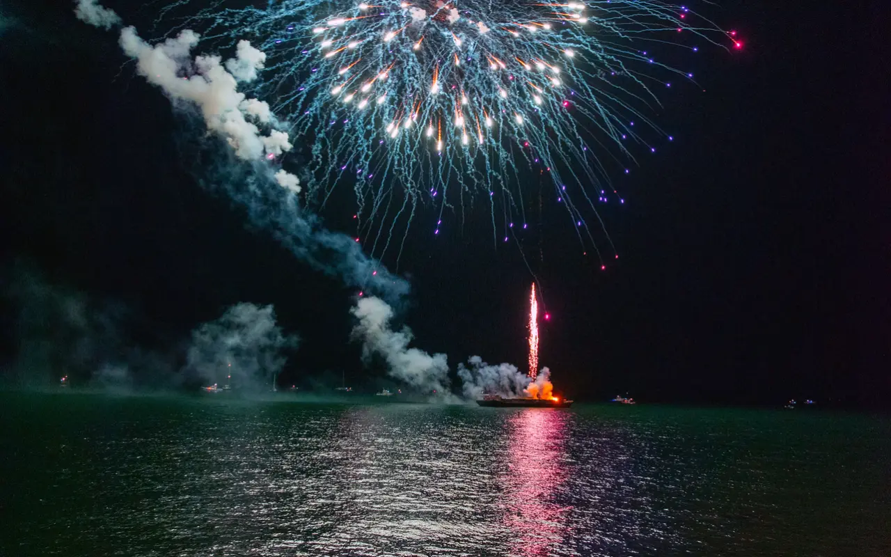 A white and blue firework over the water off Oak Bluffs at night with the lit launch barge visible on the sea below