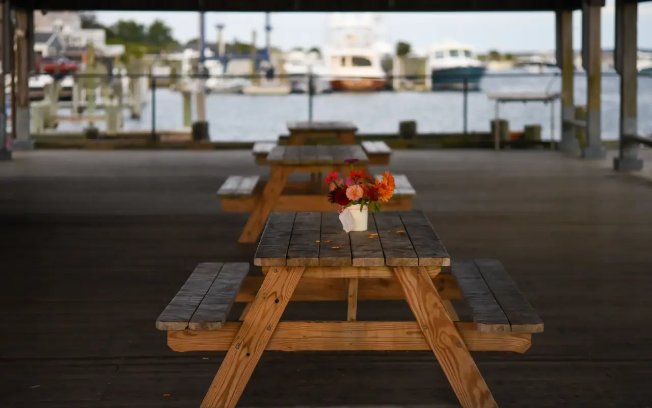 Wooden picnic tables with a jar of orange and pink flowers under a covered pier, harbor water and moored boats in the background