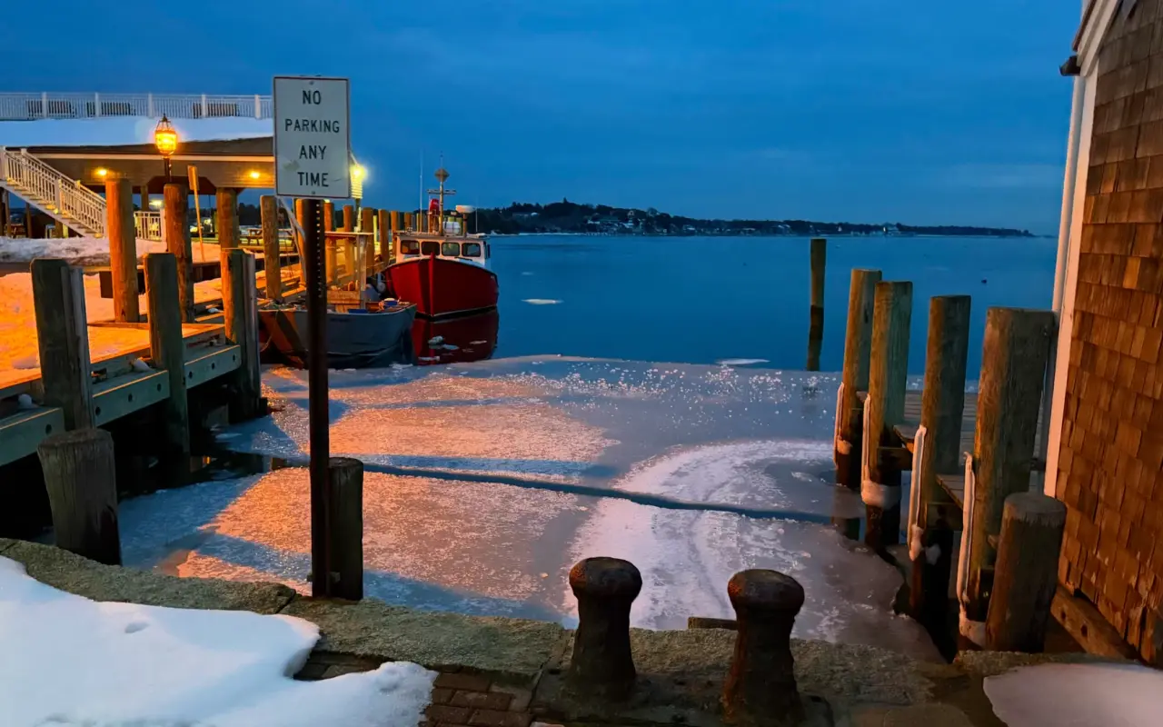 A wooden dock at Edgartown Harbor at dusk in winter with calm water and lights across the harbor
