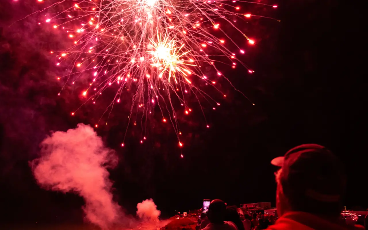 Red fireworks bursting over dark water at Oak Bluffs with a spectator silhouetted in red light in the foreground