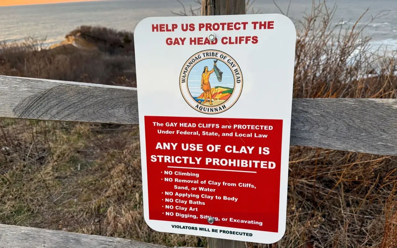 Red and white Wampanoag Tribe sign asking visitors to protect the cliffs, mounted on a wooden rail in Aquinnah
