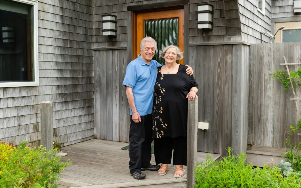 Two hosts standing at the front door of JADE House Bed and Breakfast in West Tisbury, gray shingles and a wooden door behind them