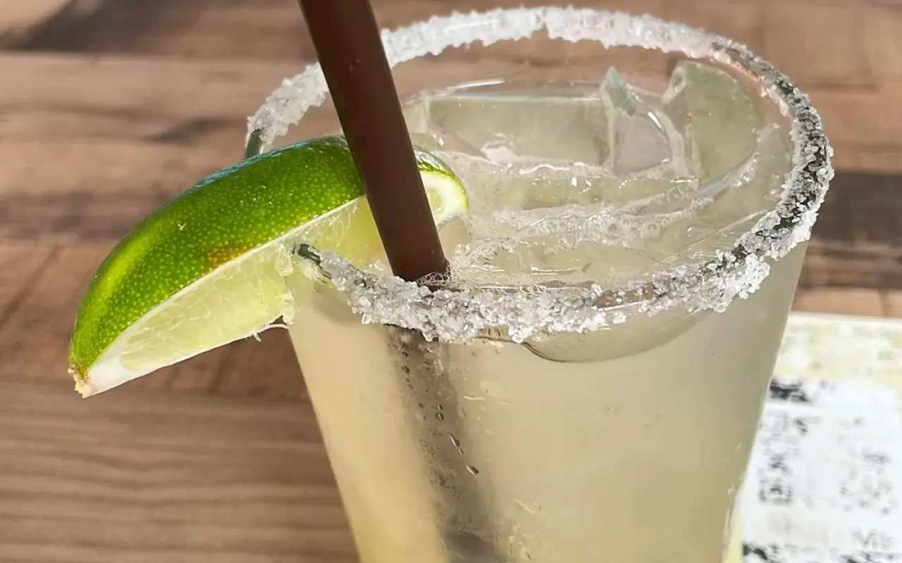 A margarita with a salted rim, a lime wedge and a straw on a wooden table