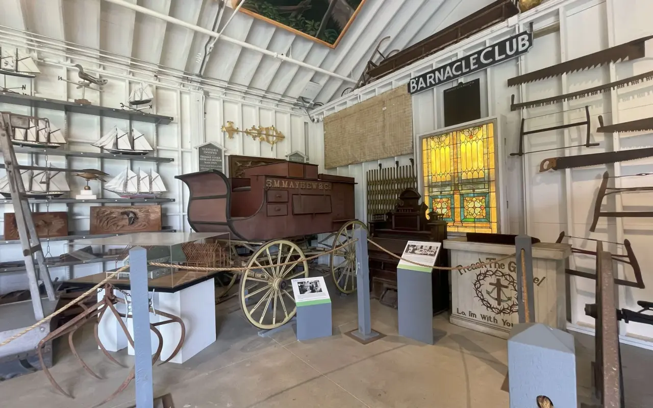 An exhibit hall in the Martha's Vineyard Museum with a horse-drawn carriage, wooden wheels and a Barnacle Club sign