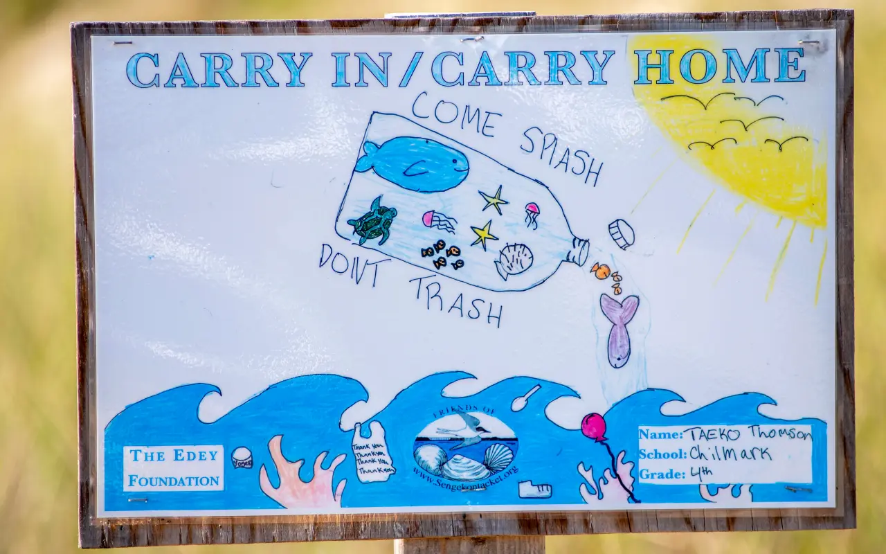 A hand-painted sign reading Carry In Carry Home with a whale, waves and a sun, posted at Menemsha