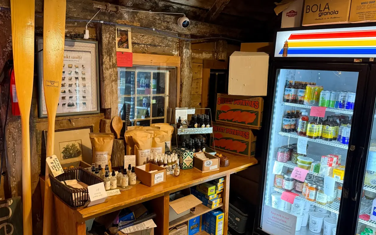 Inside a small wooden farm stand with oars on the wall, shelves of bagged and bottled goods and a glass-front refrigerator