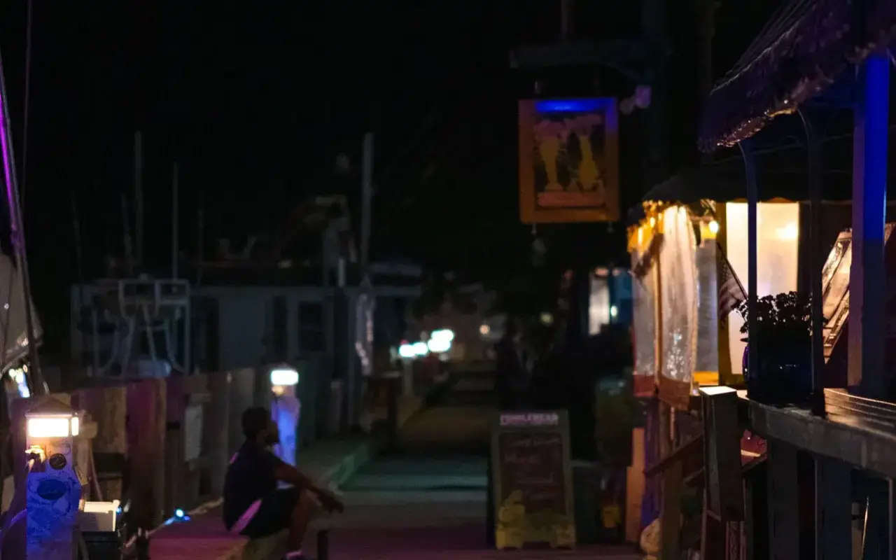 Oak Bluffs harbour boardwalk at night with lit bar signs and a person sitting on a bench