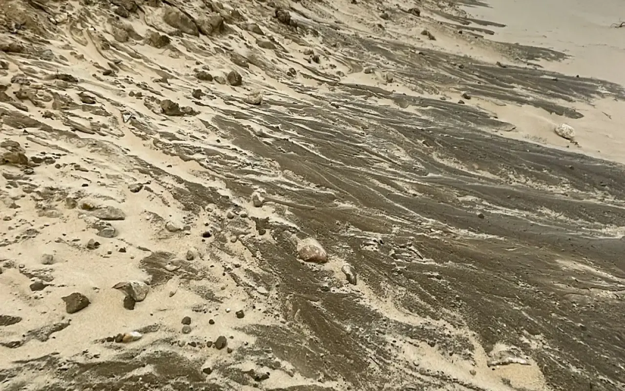 Close view of a sloping sand face streaked with dark sand and scattered pebbles at the east end of South Beach