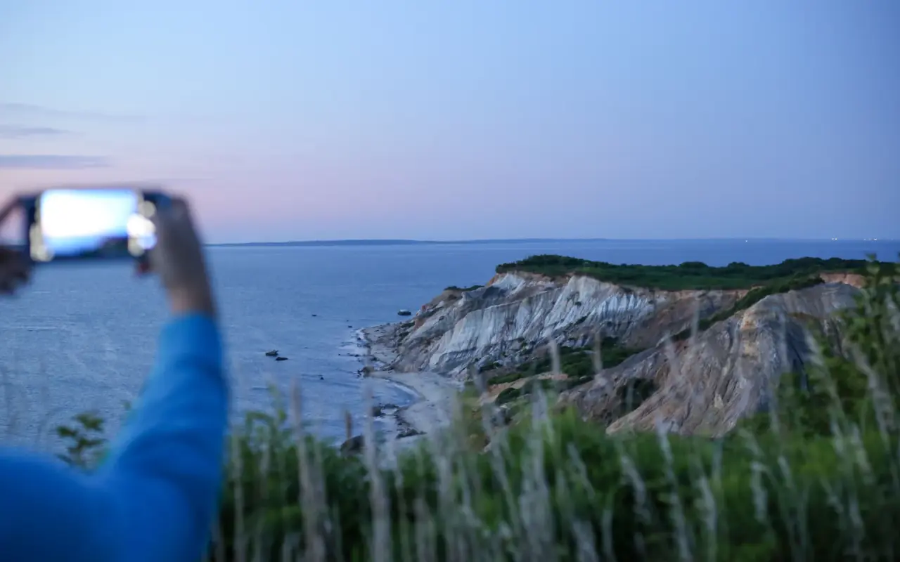 A hand holding a phone up to photograph the clay cliffs at Aquinnah at dusk