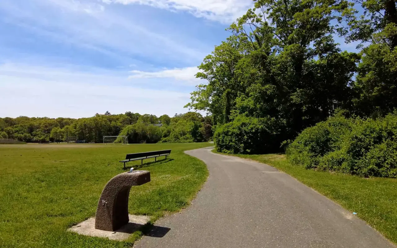 A paved path curving around open grass fields at Veterans Memorial Park in Vineyard Haven, a bench beside it and trees beyond