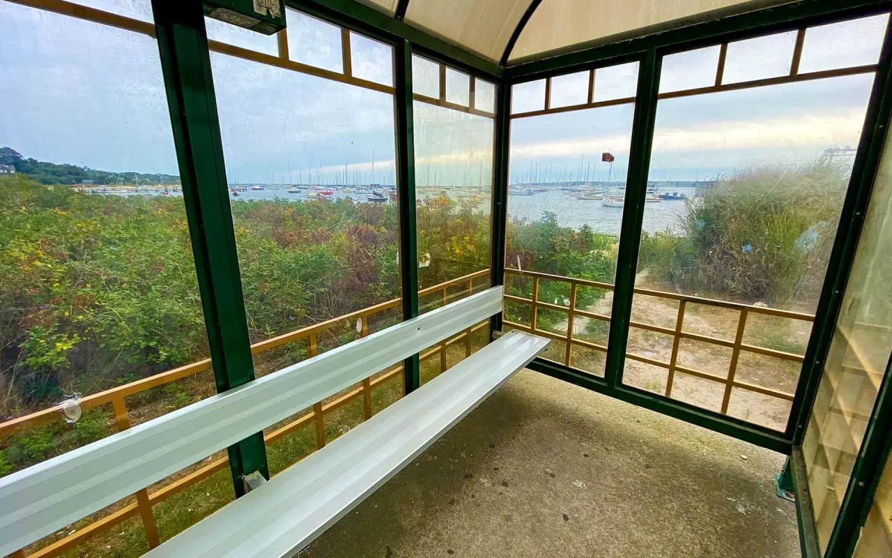 Inside a glass bus shelter in Vineyard Haven with a metal bench and a view of the harbor through the windows