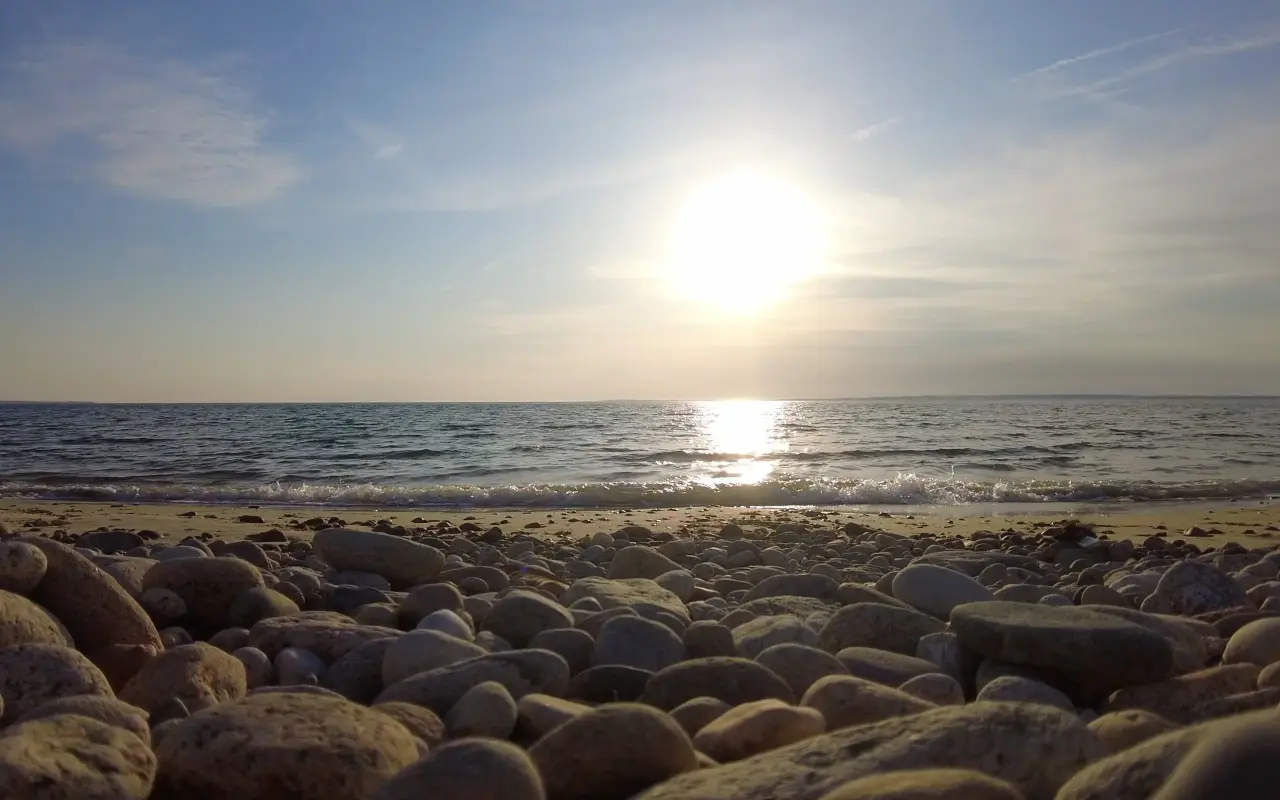 The sun low over the water from a stony beach in West Tisbury with the sky bright and the sea silver