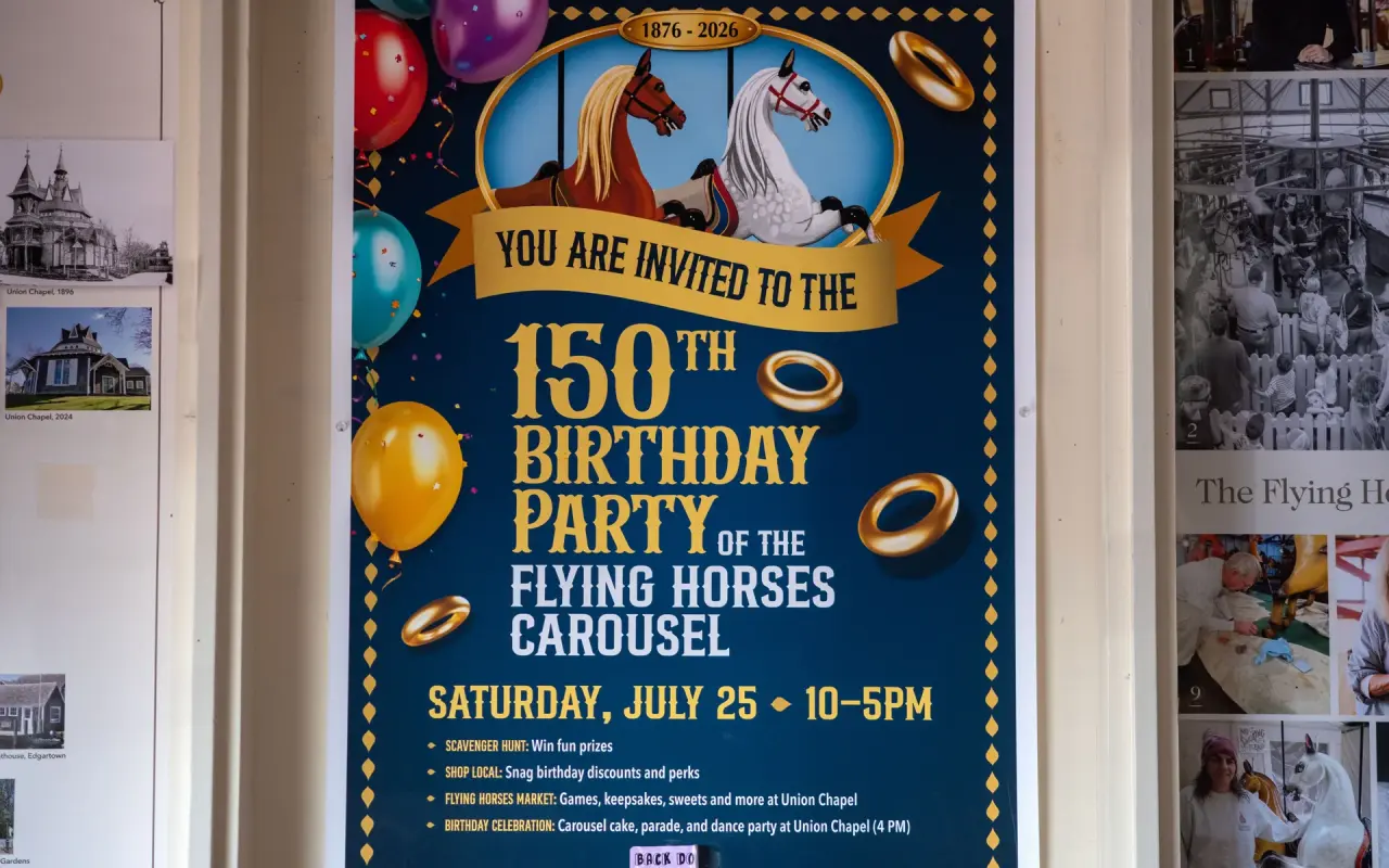 A blue and gold poster inviting visitors to the 150th birthday party of the Flying Horses Carousel, framed on a wall inside the carousel building