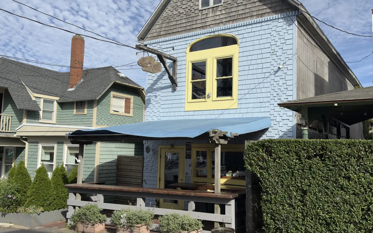 The front of Aalia's Coffee at 16 Kennebec Avenue in Oak Bluffs: a pale blue shingled building with yellow-trimmed windows and door, a carved wooden Aalia's Coffee sign on a bracket, a blue awning over a wooden counter and bench on the sidewalk, a green house next door