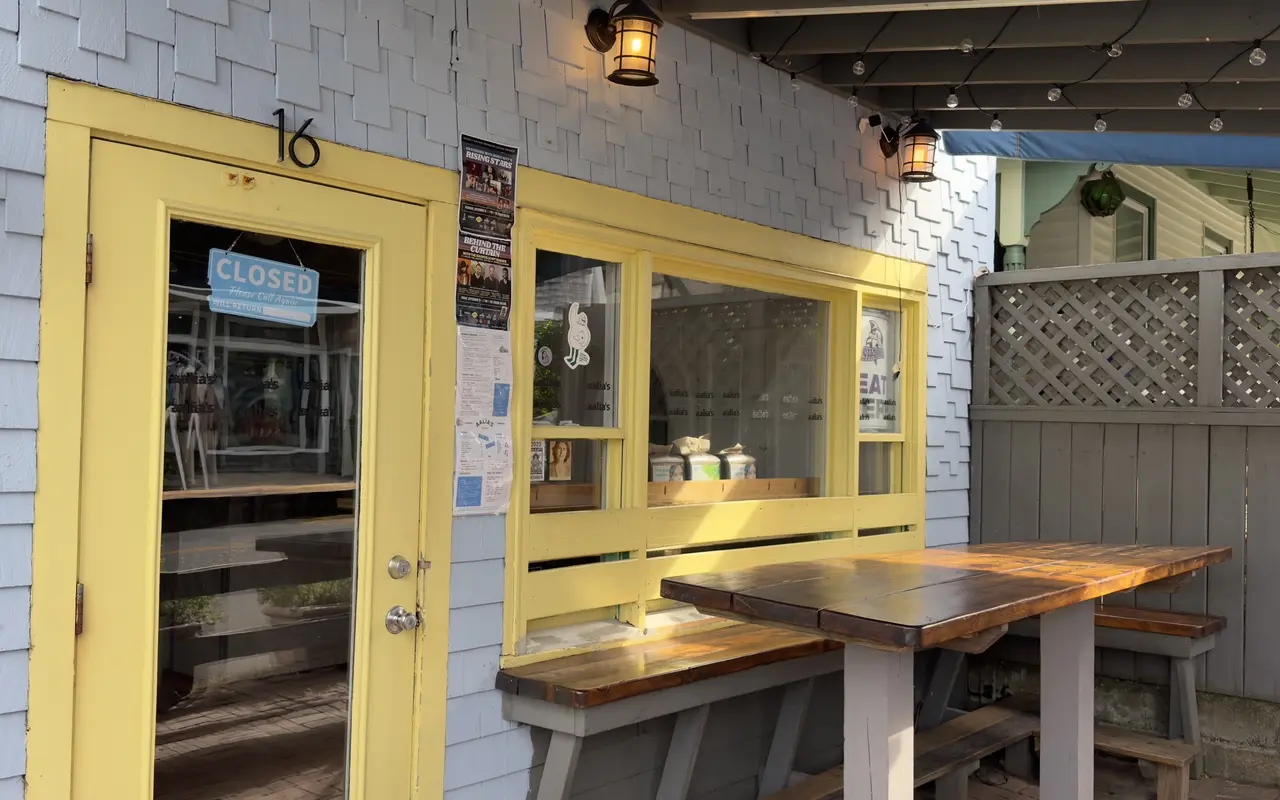 The yellow door of Aalia's Coffee in Oak Bluffs with a Closed sign, two Songwriters Festival posters taped beside it and a picnic table under string lights