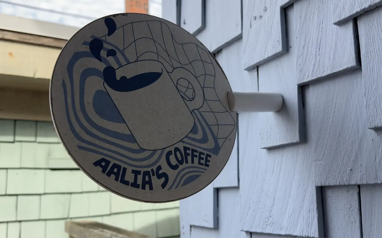Round sign with a coffee mug illustration reading Aalia’s Coffee mounted on gray shingles