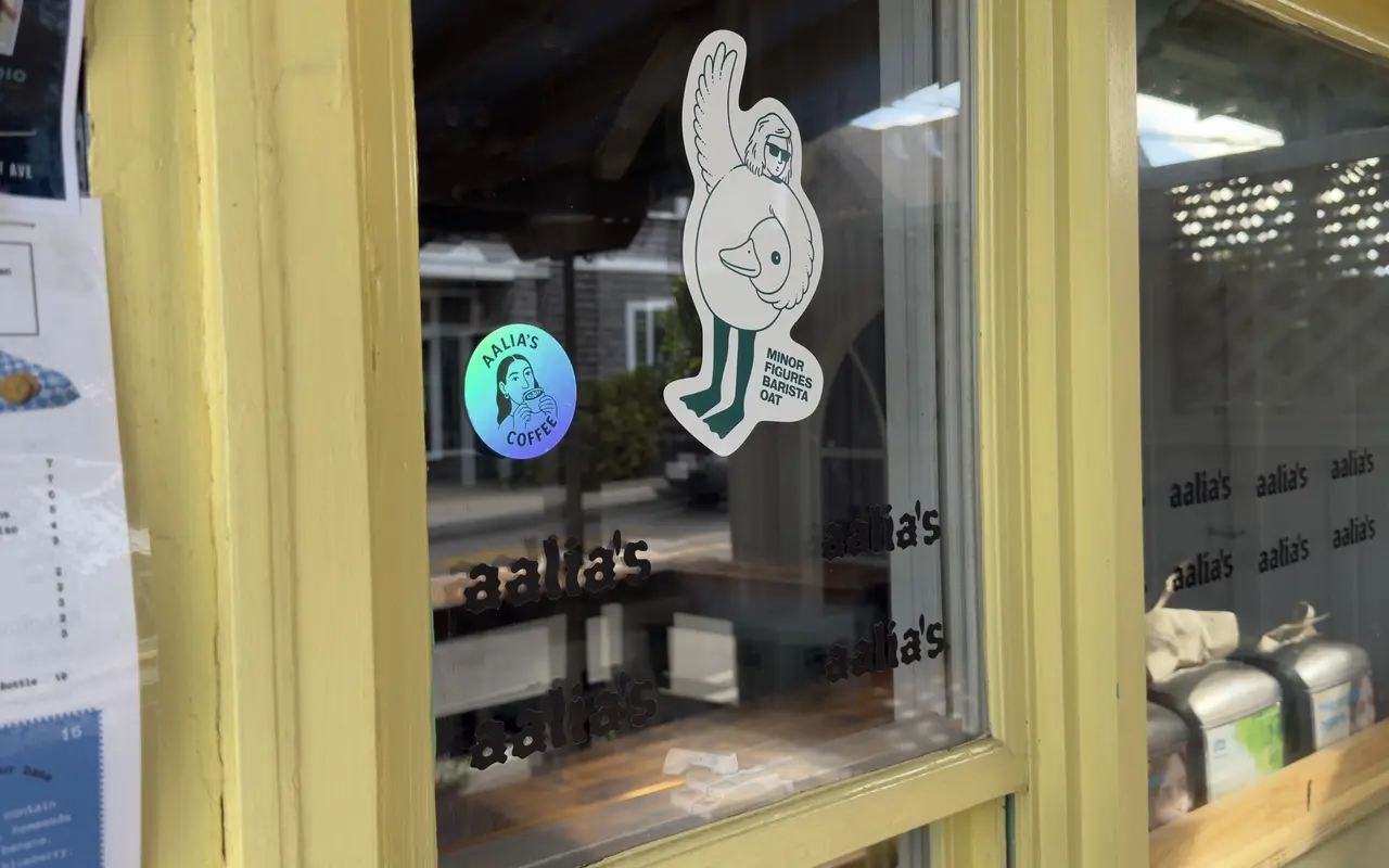 The window of Aalia's Coffee in Oak Bluffs: aalia's lettering across the glass, a holographic Aalia's Coffee sticker, a Minor Figures barista oat sticker, a printed menu of sandwiches and bowls taped to the frame, a WVVY radio flyer, and metal canisters on the counter inside