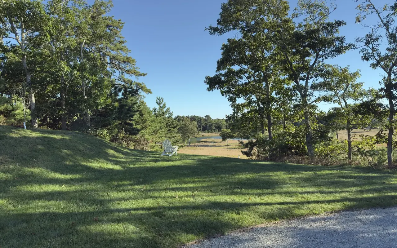 A white Adirondack chair on a green lawn between oaks and pitch pines, looking toward a tan meadow and a small pond, a gravel drive in the foreground