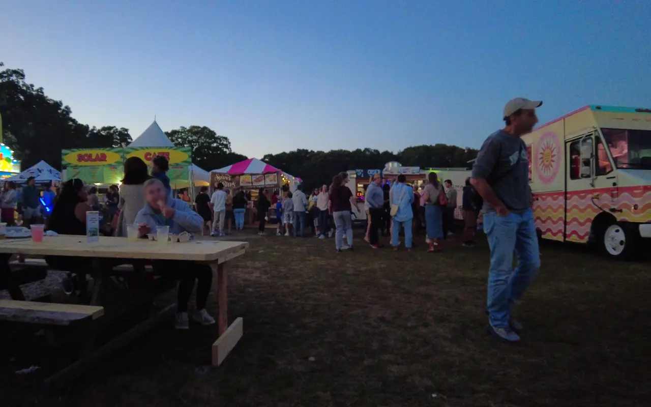 People eating at a wooden picnic table and standing in line at food trucks and tents at dusk at the Martha's Vineyard Agricultural Fair