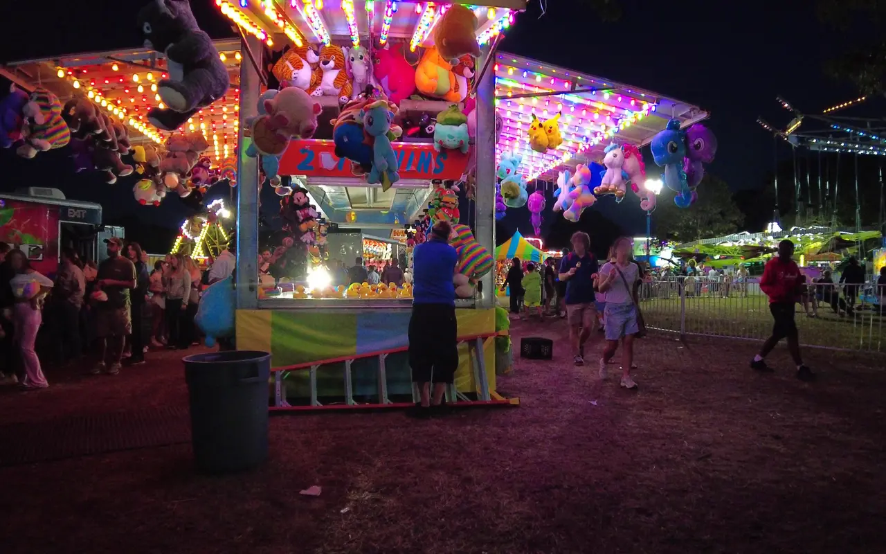 A brightly lit carnival game booth hung with plush prizes at night, people walking and lining up around it, a lit swing ride in the background