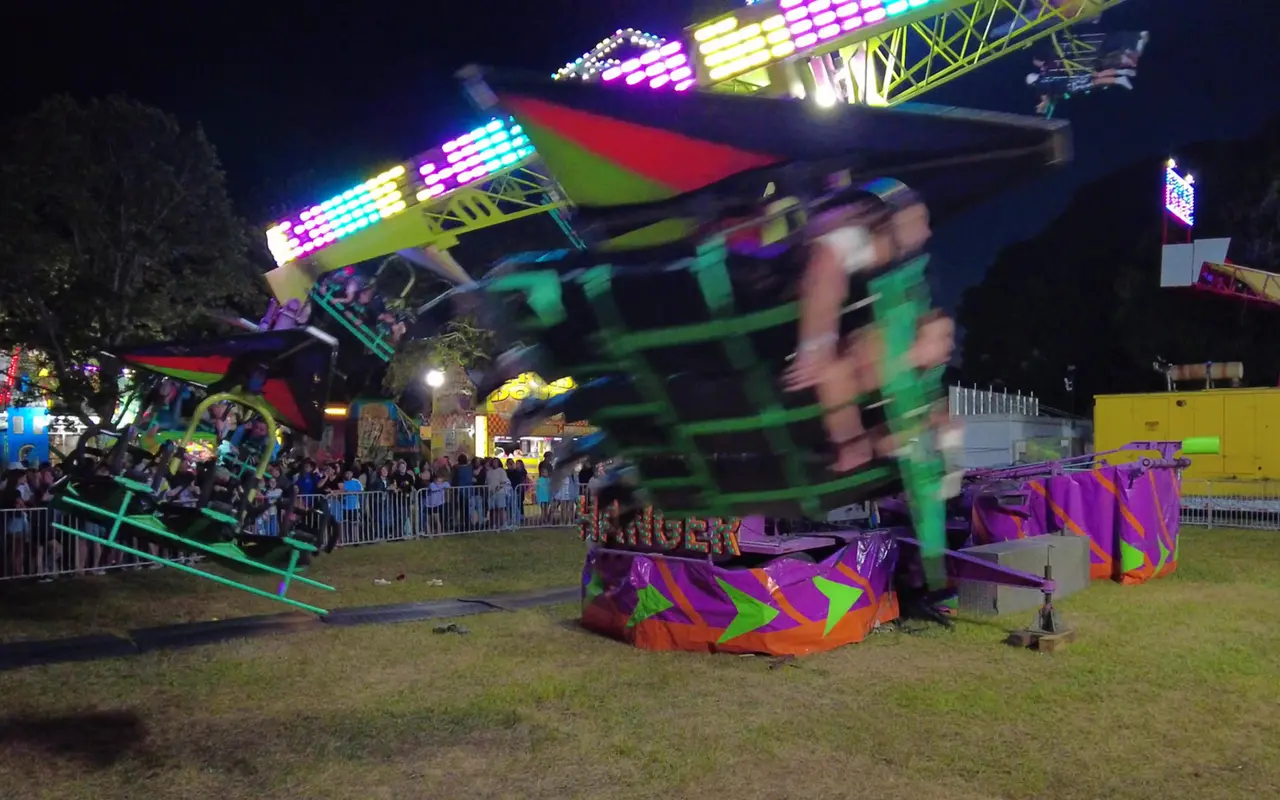 A carnival ride with neon lights swinging its riders through the air at night, people lined up along a metal fence behind