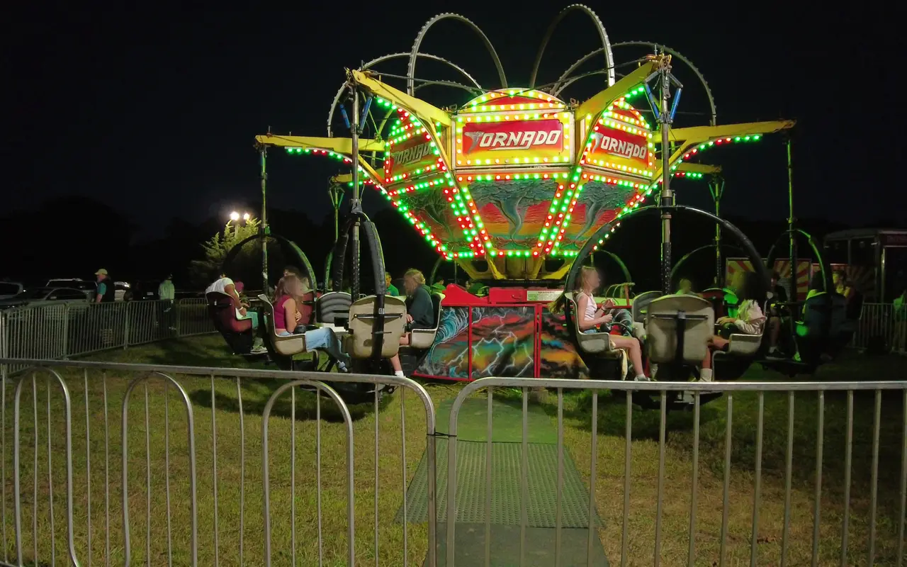 The Tornado carnival ride lit with red and green lights at night, riders in its spinning cars behind a metal fence