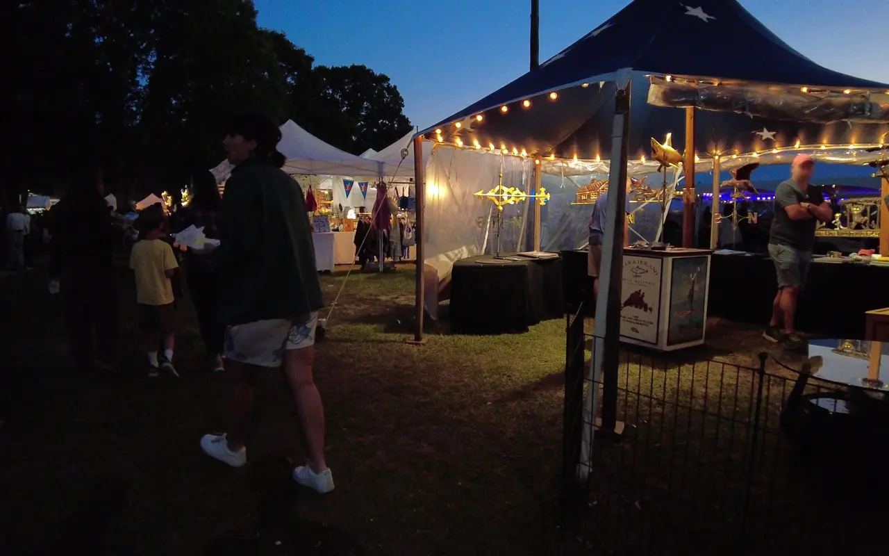 A vendor tent with string lights and gold weathervanes on display at dusk, people walking past on the grass