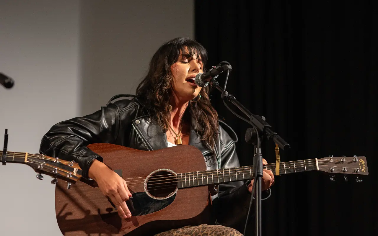 Alee singing into a microphone with an acoustic guitar on the Strand stage
