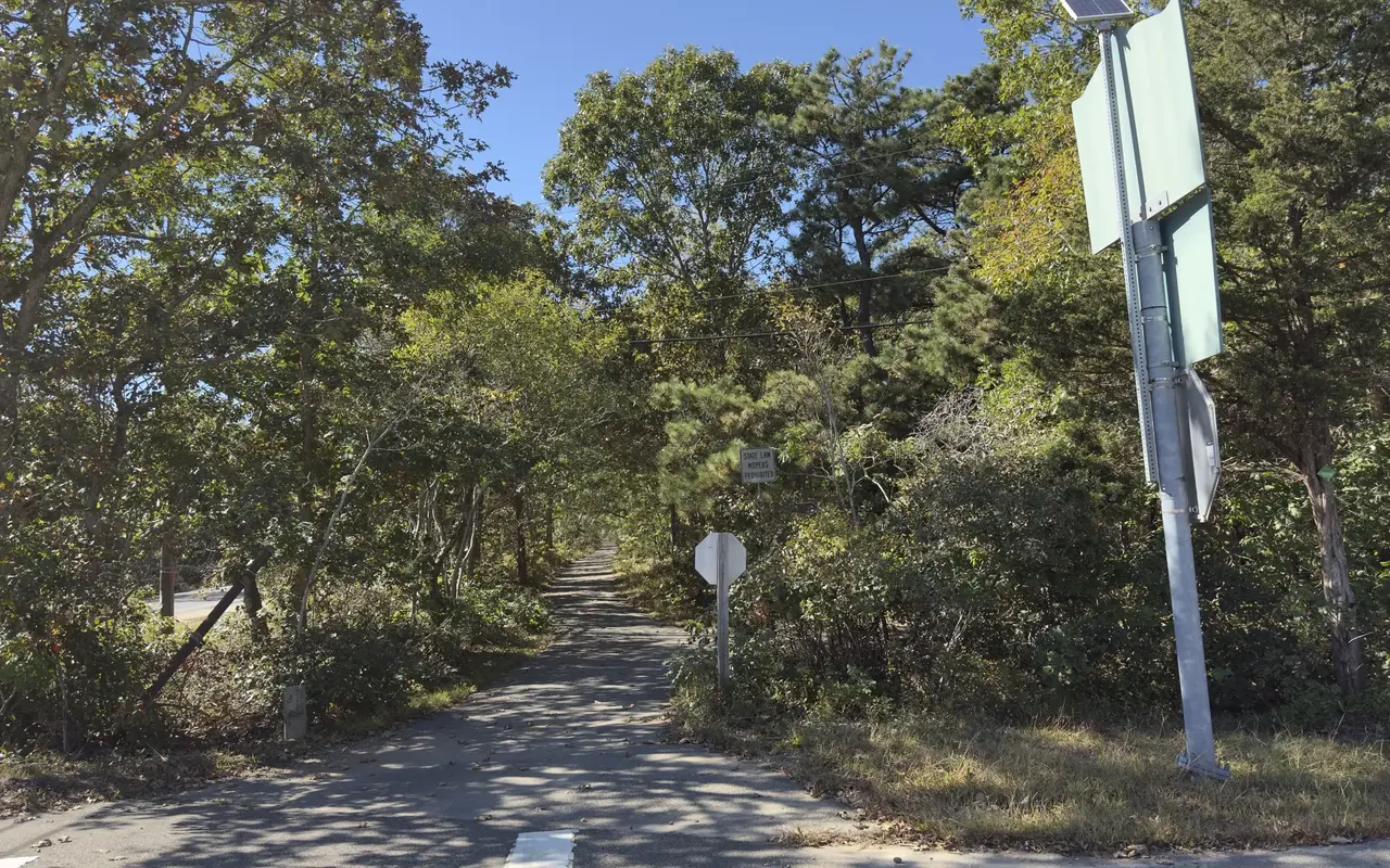 A paved bike path running straight into oak and pitch pine woods from a road crossing, with a stop sign, a sign reading State Law Mopeds Prohibited and a tall sign post on the right