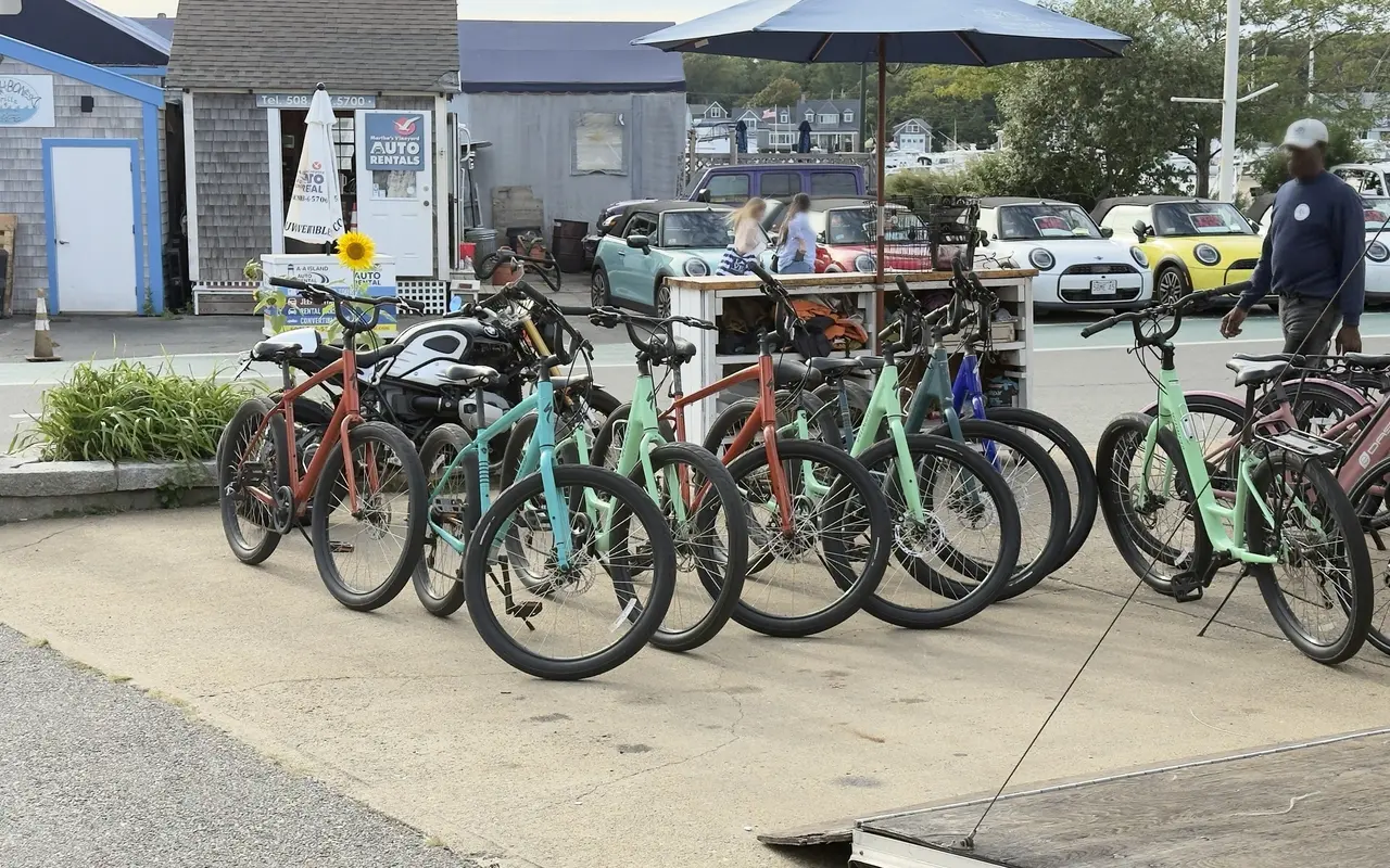 A row of rental bikes, coral, mint and teal, lined up on the pavement at a rental stand by Oak Bluffs harbor, with a rental kiosk, a Fishbones Grille sign and Mini convertibles in the lot behind