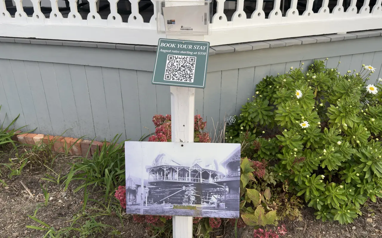 Green Book Your Stay sign with a QR code in front of a grey-blue cottage in Oak Bluffs, an old cottage photo below it