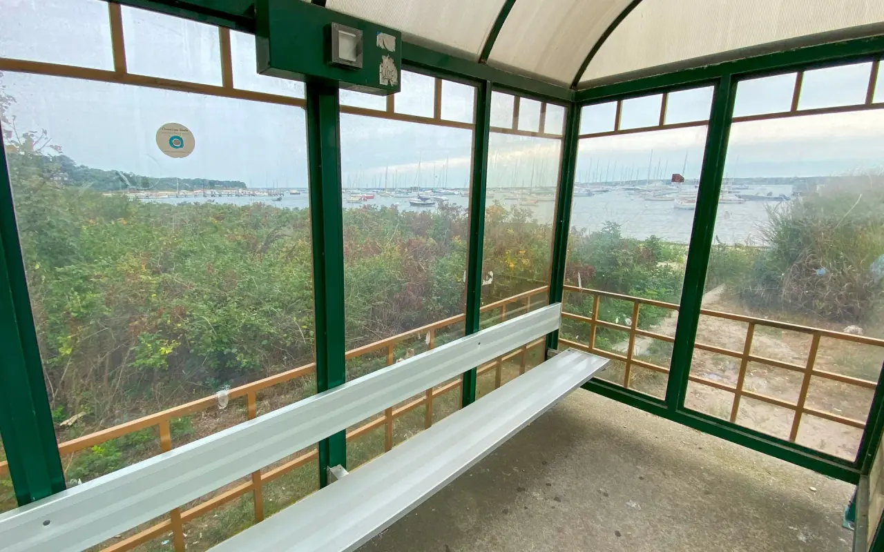 Interior of a green-framed glass bus shelter with a bench, the harbor and sailboat masts visible beyond