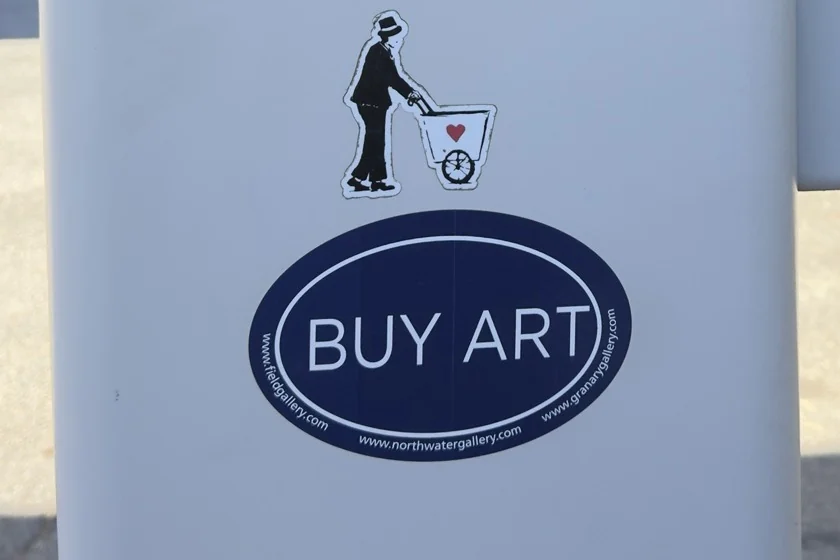 Navy oval sticker reading BUY ART with three gallery web addresses, below a small sticker of a figure pushing a cart with a heart