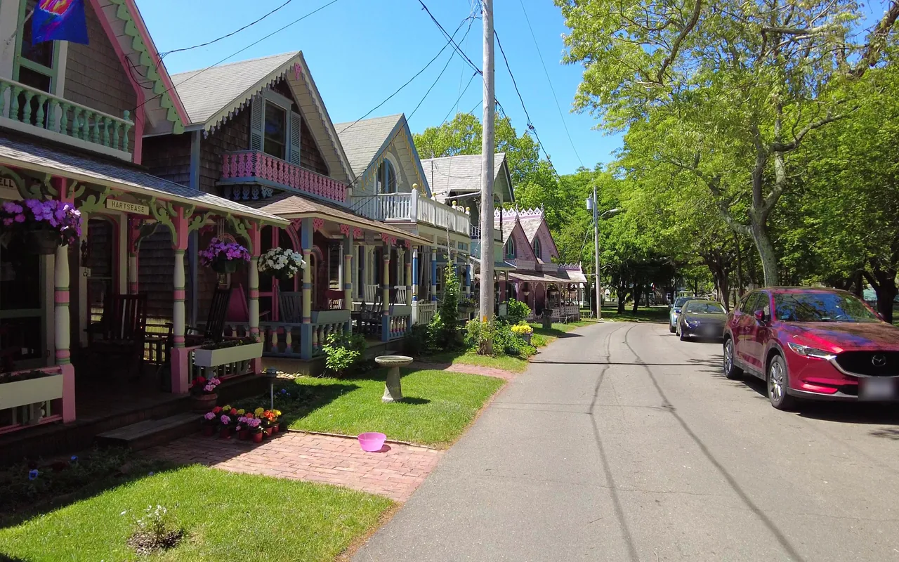 Row of colorful Victorian cottages with hanging flower baskets along a narrow paved lane, trees in leaf