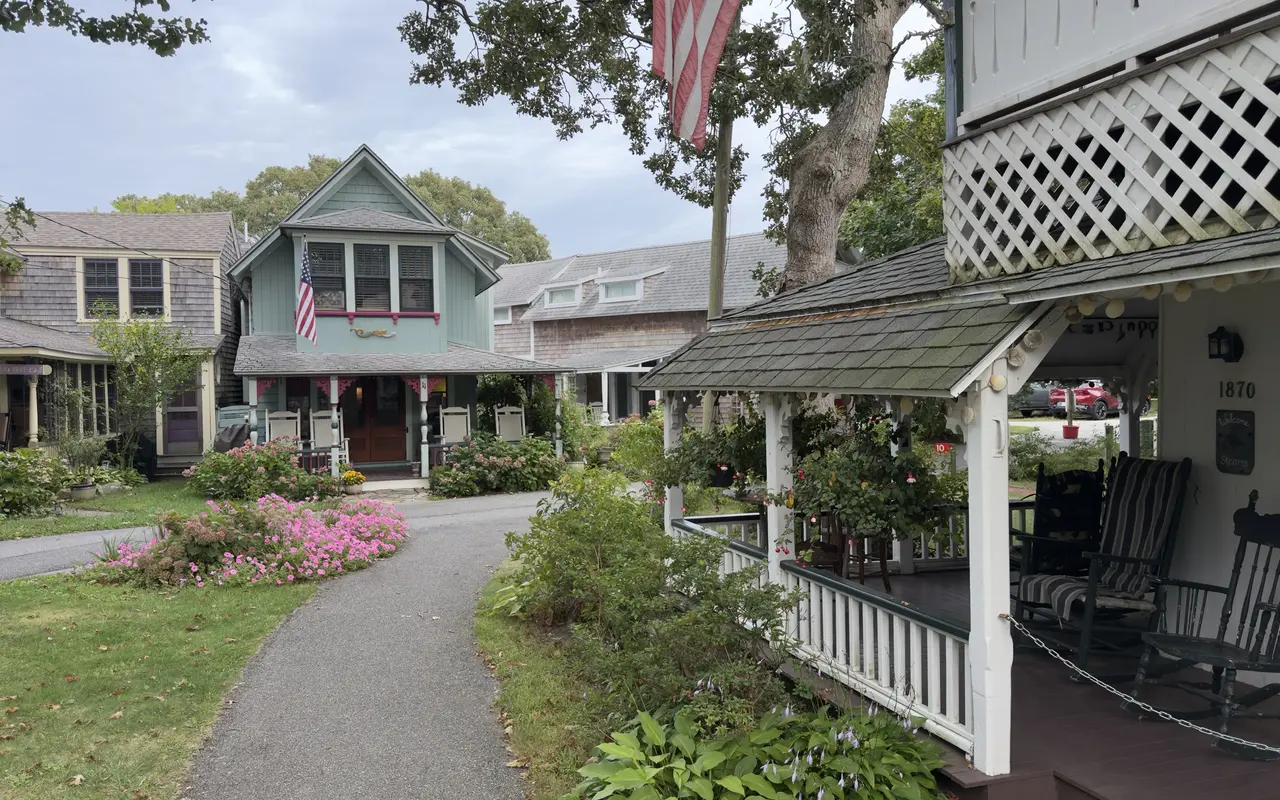 A paved lane in the Oak Bluffs Campground: a porch marked 1870 with black rocking chairs and a Welcome sign on the right, a pale blue cottage with pink trim and a flag straight ahead, pink flowers on the lawn