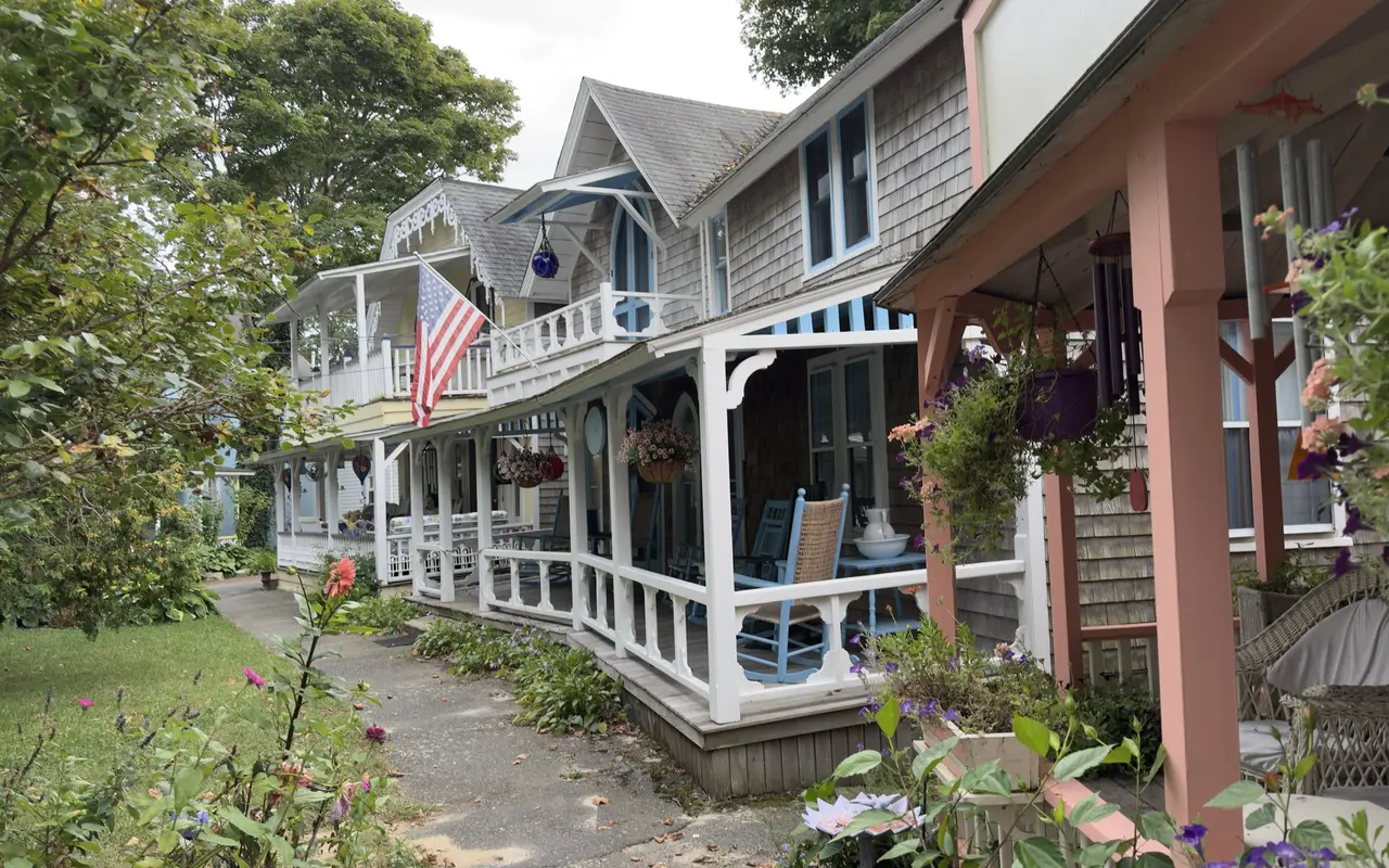 A row of cottage porches in the Oak Bluffs Campground: white railings with cutouts, blue rocking chairs, a white pitcher and bowl on a table, hanging baskets, an American flag on the balcony above and a coral-painted porch with wicker chairs in the foreground