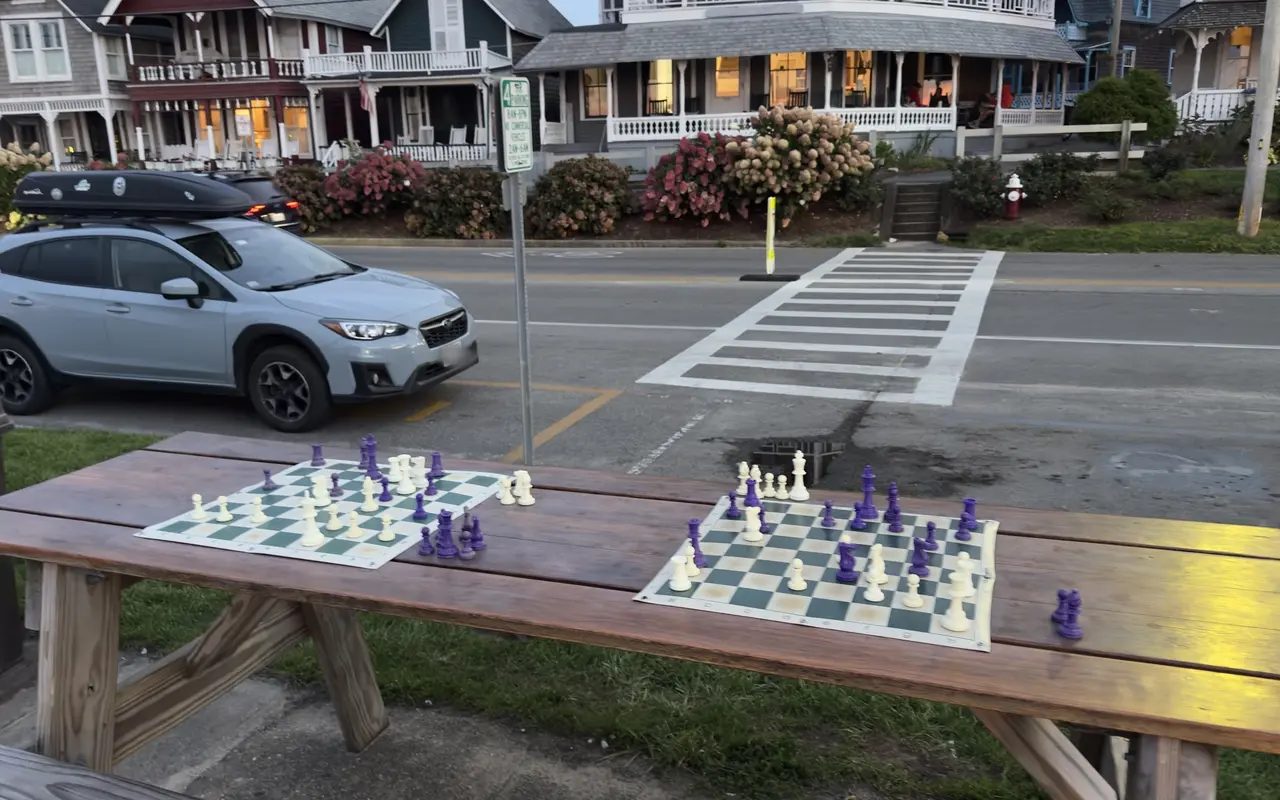 Two chess boards with games in progress on a wooden picnic table at dusk in Oak Bluffs, a parked car and Victorian cottages across the street