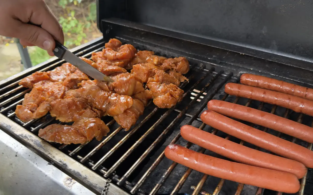 Marinated chicken pieces and six hot dogs cooking on a gas grill, a hand turning the chicken with a knife