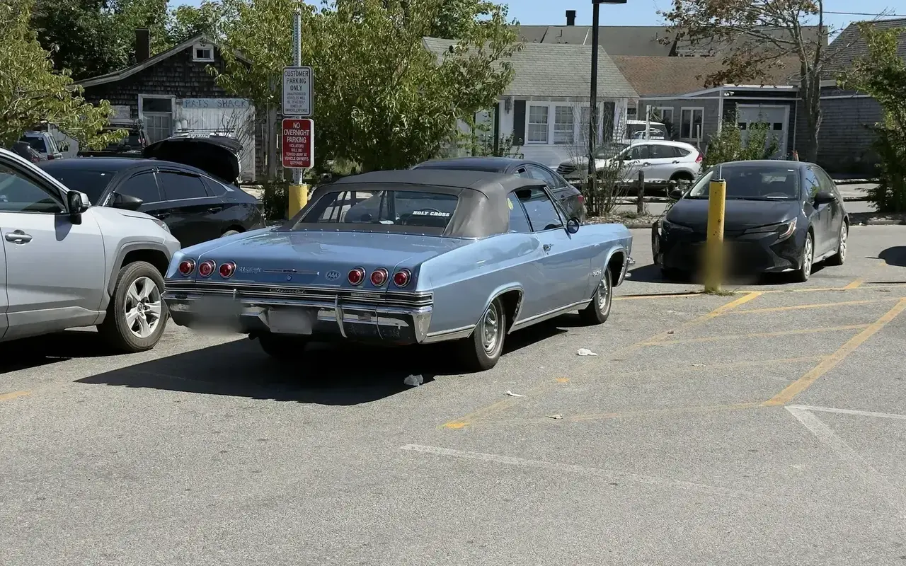 A light blue mid-1960s Chevrolet Impala convertible with a black top parked in a Vineyard Haven lot