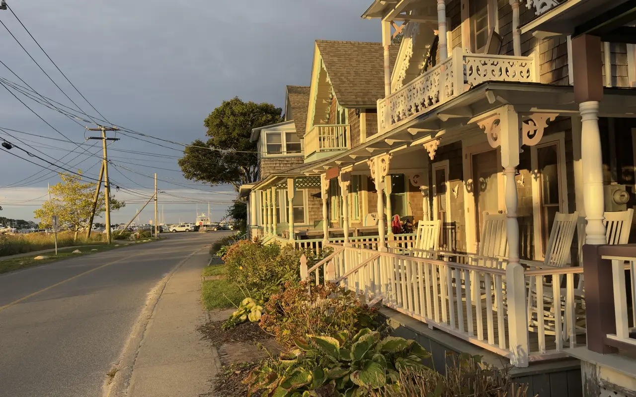 A row of shingled cottages with cream scrollwork balconies and white rocking chairs on the harbor side of Oak Bluffs at golden hour, masts visible at the end of the road