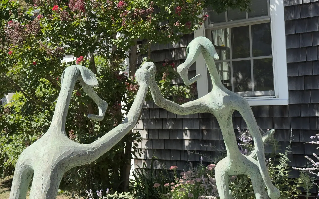 Close-up of two green-patina sculpted figures holding hands and dancing in a front garden in Vineyard Haven, a crape myrtle in bloom, Russian sage and a grey-shingled wall with a white window behind them